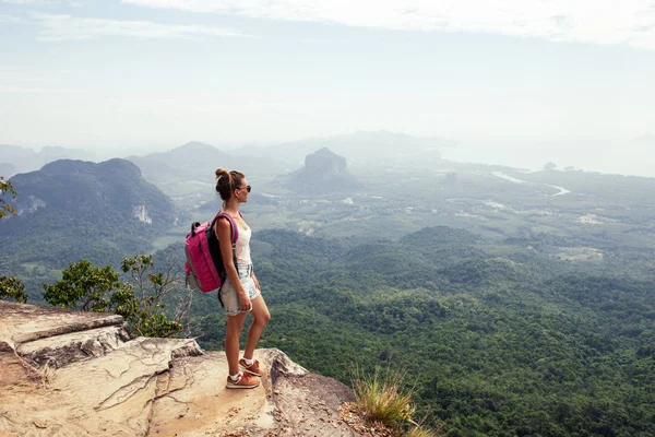 Young woman traveler with backpack standing on top of a mountain and enjoying the view