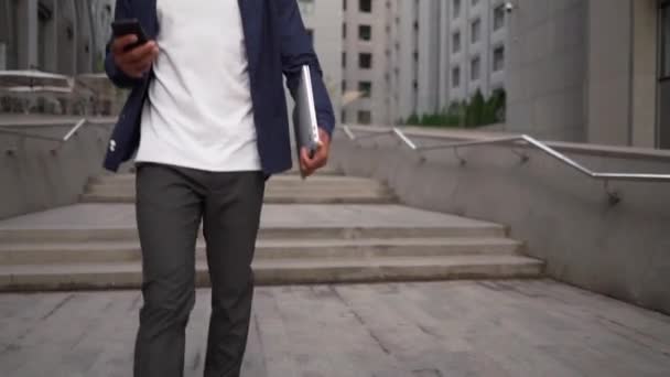 Etniczny człowiek z laptopem i smartfonem spacerujący ulicą — Wideo stockowe