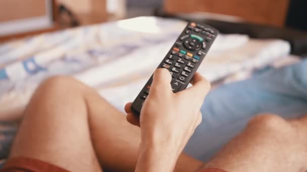 Hombre cansado se acuesta en la cama y emocionalmente mira la televisión con control remoto — Vídeos de Stock