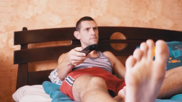Втомлений чоловік обманює на ліжку й емоційно дивиться телевізор з дистанційним контролем — стокове відео