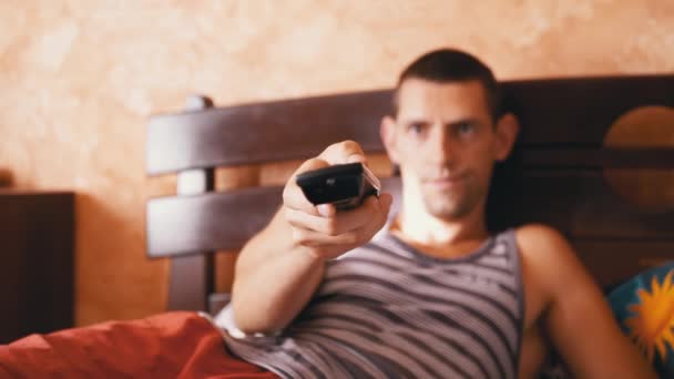 Homem Cansado jaz na cama e emocionalmente assiste TV com controle remoto — Vídeo de Stock