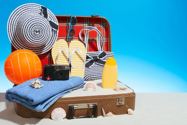 Nice opened suitcase full of white sand and various stuff for beach trip standing on blue background