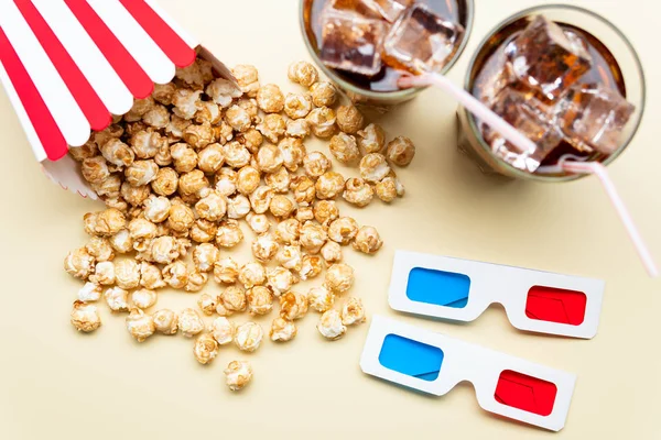 Carton of sweet popcorn and two glasses of tasty cold beverage near 3D glasses on light yellow background