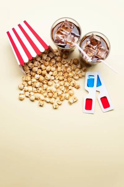 Carton of sweet popcorn and two glasses of tasty cold beverage near 3D glasses on light yellow background