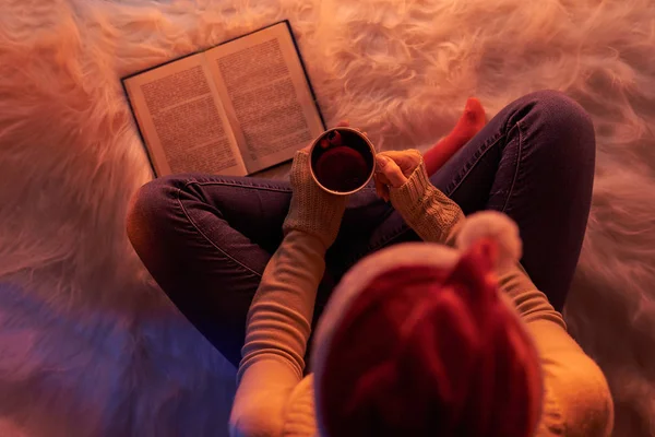 From above shot of woman in Santa hat sitting with mug on white fur in soft light reading book