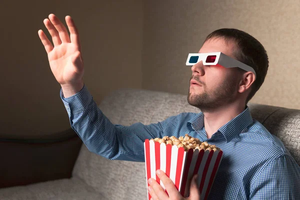 A grown man wearing 3D glasses waving his hand and eating popcorn, while sitting on a comfortable sofa at home