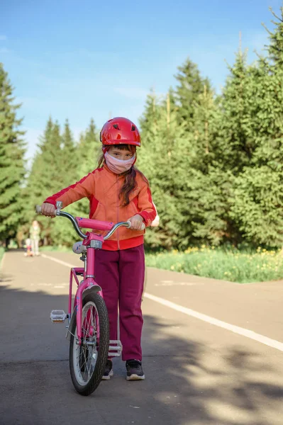 Bambina in maschera protettiva cavalca una bicicletta in un parco cittadino . — Foto Stock
