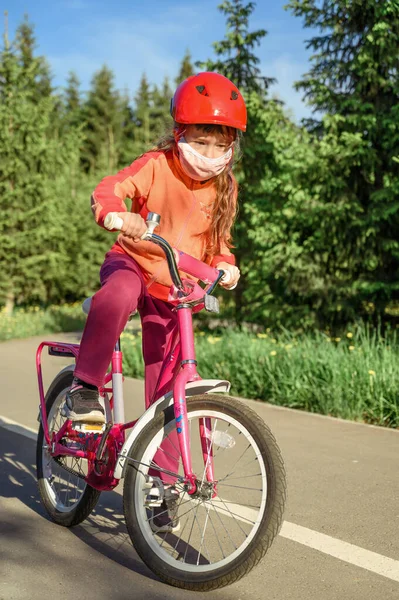 Bambina in maschera protettiva cavalca una bicicletta in un parco cittadino . — Foto Stock