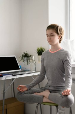 Genç kız evde eğitim görüyor, yoga egzersizleri yapıyor, lotus pozisyonunda oturuyor, meditasyon yapıyor..