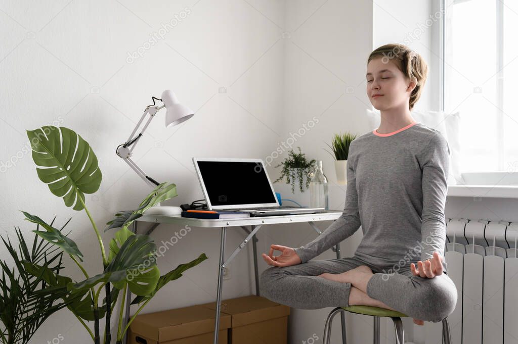 Teen girl is training at home, doing yoga exercises, sitting in lotus position, meditation.