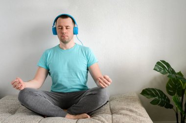 Genç adam koltukta nilüfer pozisyonunda oturur, kulaklıkla müzik dinler. Evde yoga..