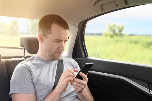 Jovem passageiro do sexo masculino em um carro detém um smartphone, escreve uma mensagem em redes sociais . — Fotografia de Stock