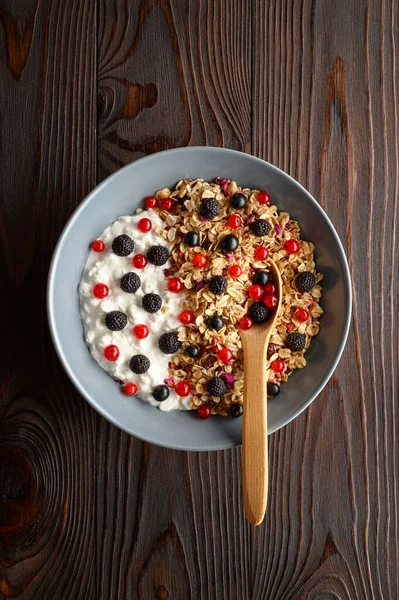 Tigela com granola de aveia, iogurte e framboesas pretas, groselhas pretas e vermelhas em um fundo de madeira marrom . — Fotografia de Stock