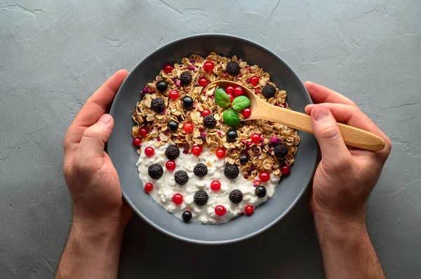 Mãos masculinas estão segurando uma colher de madeira e uma tigela com granola de aveia, iogurte e framboesas pretas, groselhas pretas e vermelhas . — Fotografia de Stock