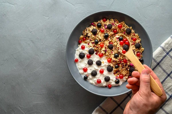 A mans hand holds a wooden spoon over a bowl with oat granola, yogurt and black raspberries, black and red currants. — Stock Photo, Image