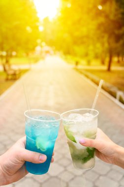 Bir şehir parkında erkek ve kadın elleri buzlu meyve limonatasıyla plastik bardakları tutuyor. Alkolsüz kokteylleri tazeliyorum. Yaz güneşli bir gün.