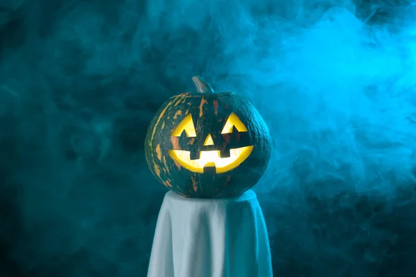 A funny ghost with the head of a glowing jack-o-lantern pumpkin stands in the fog at dusk.