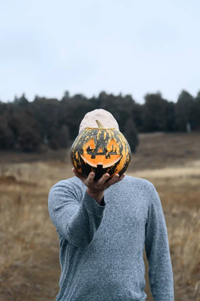 Homem Anônimo em uma camisola segura uma abóbora de Halloween esculpida na frente de seu rosto ao ar livre. Conceito sem rosto. — Fotografia de Stock