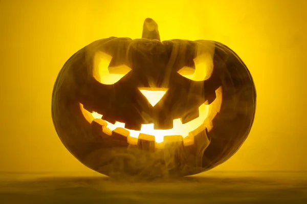 Enge gloeiende pompoen Jack-o-lantaarn in de rook op een gele achtergrond. — Stockfoto