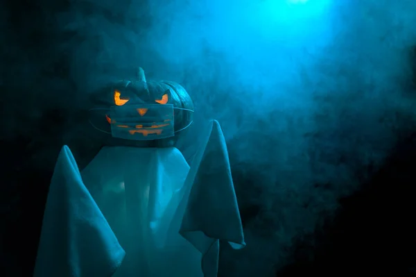 Símbolo de Halloween. Um fantasma com uma cabeça assustadora de uma abóbora luminosa Jack-o-Lanterna em uma máscara médica protetora fica no nevoeiro ao entardecer. Novo conceito normal. — Fotografia de Stock