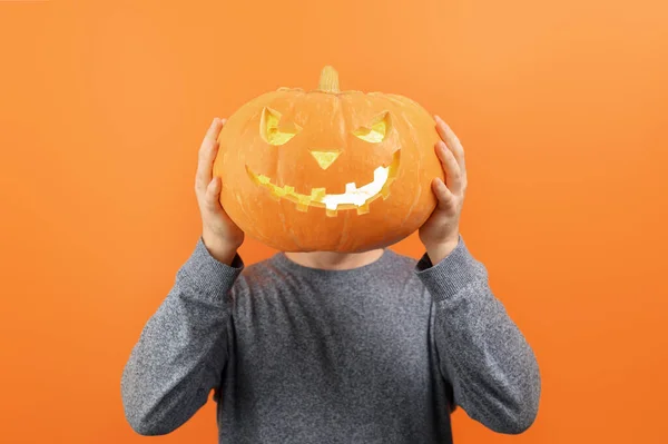 Anonymous Man in a sweater holds a carved Halloween pumpkin in front of his face on a orange background.
