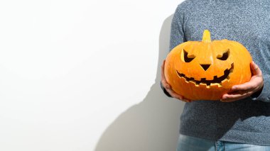 Tanınmayan bir adam önünde oyulmuş bir Cadılar Bayramı kabağı tutuyor. Beyaz arka planda turuncu bir balkabağı olan mavi kazaklı bir adam..