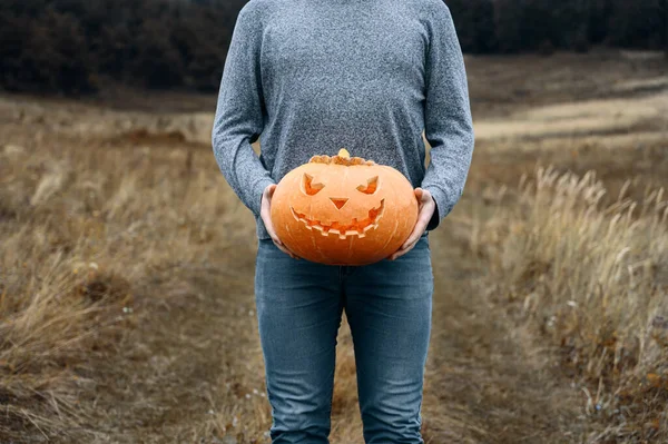 Hombre anónimo en ropa azul sostiene una calabaza de Halloween naranja tallada en frente de él, al aire libre. — Foto de Stock