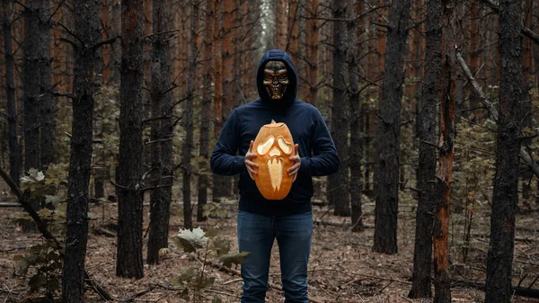 Um homem com uma máscara de crânio segura na frente dele uma abóbora de Halloween brilhante em uma floresta escura. — Fotografia de Stock