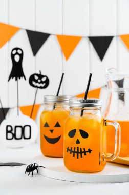 Cadılar Bayramı balkabağı buzlu kokteylleri cam kavanozlarda beyaz bir masada korkunç yüzlerle süslenmiş. Duvarda DIY Cadılar Bayramı Partisi dekorasyonu.