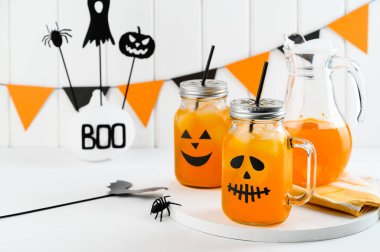 Cadılar Bayramı balkabağı buzlu kokteylleri cam kavanozlarda beyaz bir masada korkunç yüzlerle süslenmiş. Duvarda DIY Cadılar Bayramı Partisi dekorasyonu.