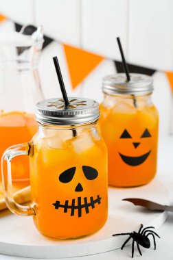 Cadılar Bayramı balkabağı buzlu kokteylleri cam kavanozlarda beyaz bir masada korkunç yüzlerle süslenmiş. Duvarda DIY Cadılar Bayramı Partisi dekorasyonu.