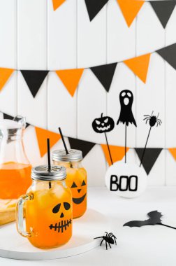 Cadılar Bayramı balkabağı buzlu kokteylleri cam kavanozlarda beyaz bir masada korkunç yüzlerle süslenmiş. Duvarda DIY Cadılar Bayramı Partisi dekorasyonu.