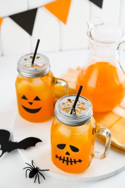 Cadılar Bayramı balkabağı buzlu kokteylleri cam kavanozlarda beyaz bir masada korkunç yüzlerle süslenmiş. Duvarda DIY Cadılar Bayramı Partisi dekorasyonu.