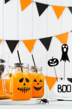 Cadılar Bayramı balkabağı buzlu kokteylleri cam kavanozlarda beyaz bir masada korkunç yüzlerle süslenmiş. Duvarda DIY Cadılar Bayramı Partisi dekorasyonu.
