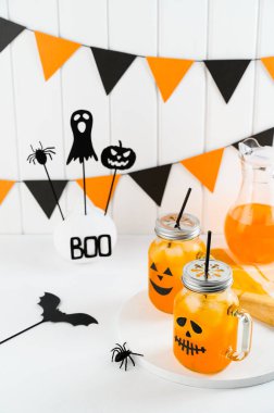 Cadılar Bayramı balkabağı buzlu kokteylleri cam kavanozlarda beyaz bir masada korkunç yüzlerle süslenmiş. Duvarda DIY Cadılar Bayramı Partisi dekorasyonu.