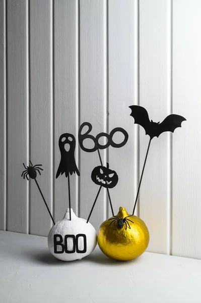 Halloween home decorations. Painted white and gold pumpkins with scary black Halloween objects on a white wooden wall background.