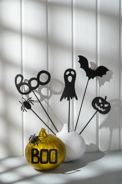 Halloween home decorations. Painted white and gold pumpkins with scary black Halloween objects on a white wooden wall background.