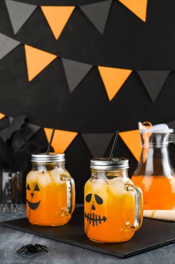 Cadılar Bayramı balkabağı buzlu kokteylleri cam kavanozlarda karanlık bir masada korkunç yüzlerle süslenmiş. DIY Cadılar Bayramı Partisi dekorasyonu siyah duvarda.