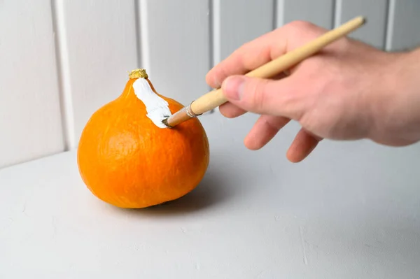 Concepto de Halloween. Una mano masculina con un pincel pinta una calabaza naranja blanca sobre la mesa. — Foto de Stock