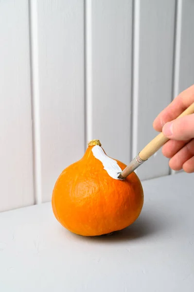 Halloween-Konzept. Eine männliche Hand mit dem Pinsel malt einen orangefarbenen Kürbis weiß auf den Tisch. — Stockfoto