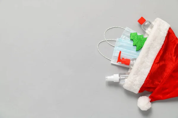 Sağlık maskesi, dezenfektan şişesi, Noel Baba şapkası ve açık gri arka planda Noel süslemeleri..