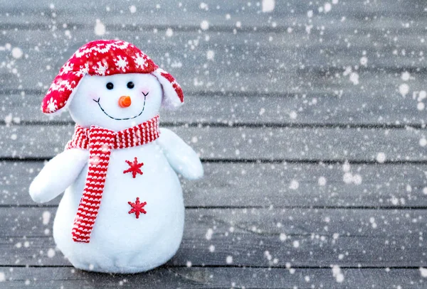 cheerful and funny snowman in a red hat and red scarf on a gray wooden background. falling snow texture