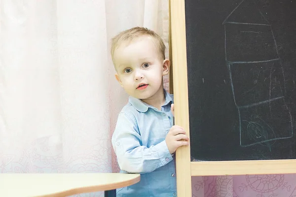 Desenvolvimento infantil precoce. Aulas preparatórias para o jardim de infância. Menino loiro bonito com uma camisa azul jeans está ao lado de uma placa escolar preta — Fotografia de Stock