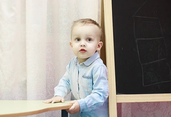 Desenvolvimento infantil precoce. Aulas preparatórias para o jardim de infância. Menino loiro bonito com uma camisa azul jeans está ao lado de uma placa escolar preta — Fotografia de Stock