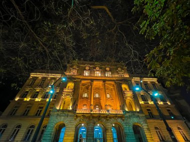 Budapeşte Üniversitesi binasının dış cephesinde ağaçlarla çevrili sarı ve mavi ışıklar olan bir gece çekimi..