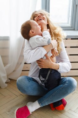 Küçük tatlı bir oğlu olan genç bir anne pencerenin yanındaki parlak, rahat bir oturma odasında eğleniyor.