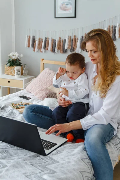 一位年轻的母亲带着心爱的儿子躺在床上 带着笔记本电脑 具有儿童概念的家庭工作 — 图库照片
