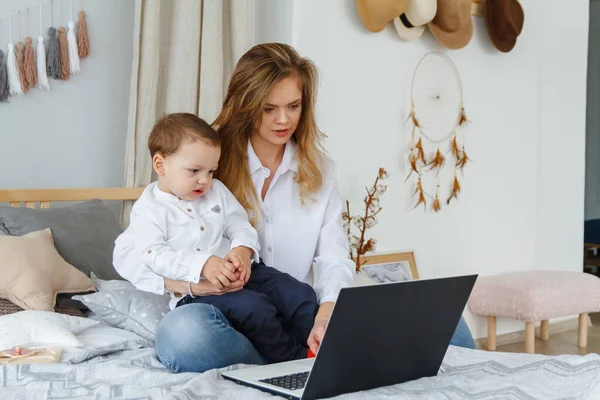 A young mother with her beloved son in the bedroom on the bed with a laptop. Work at home with a child concept
