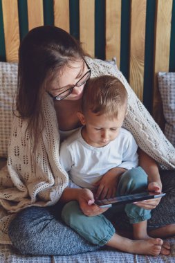 Yatakta battaniyenin altında bir tabletle genç anne ve oğlu.