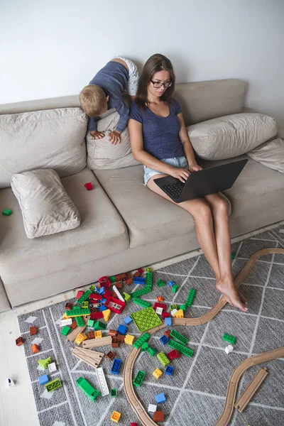 Niño juega en el sofá, mientras que la mamá trabaja con el ordenador portátil, distrae y — Foto de Stock